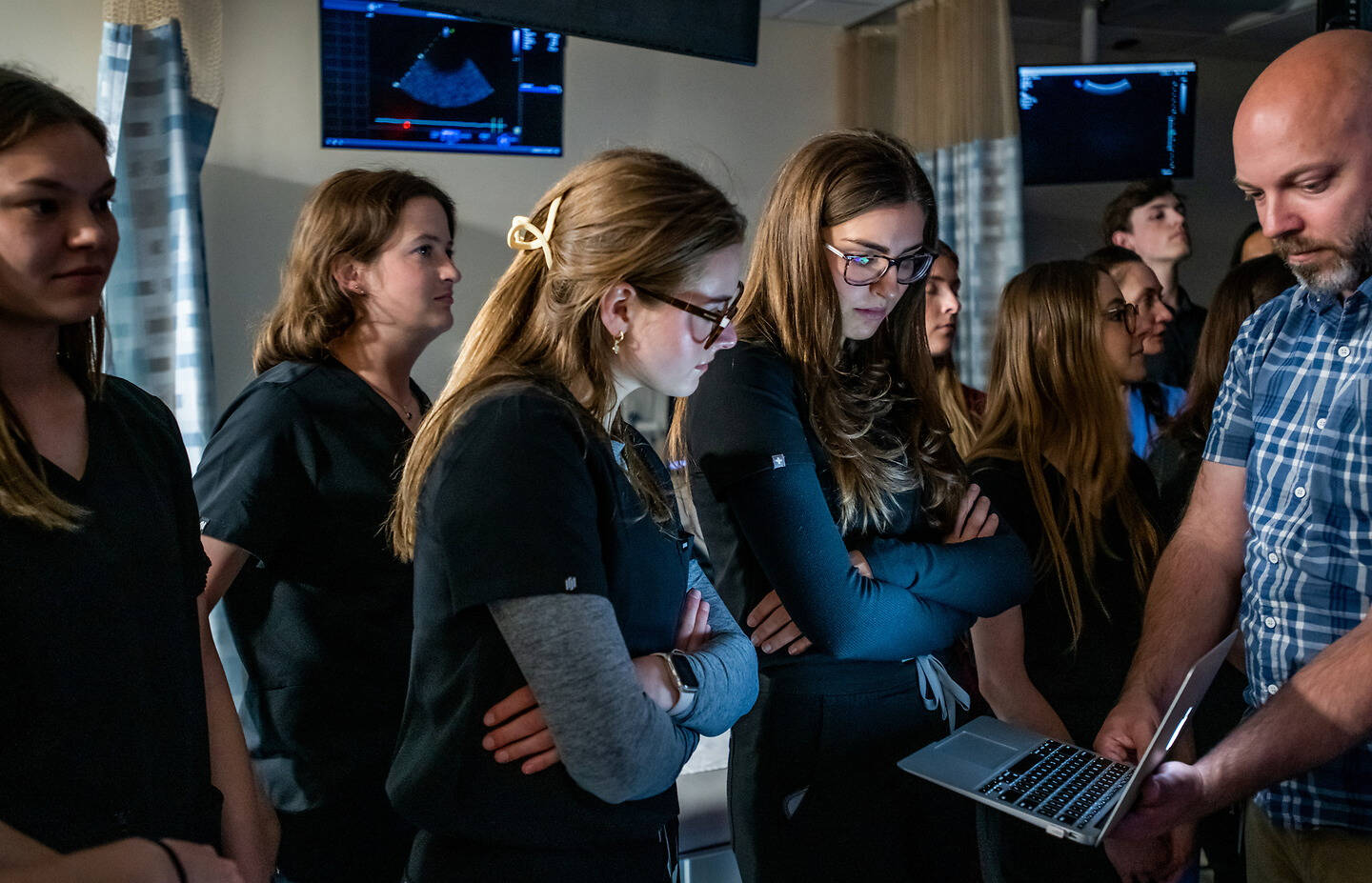 A group of individuals in medical attire gather around a person holding a laptop, attentively observing ultrasound images displayed on screens.